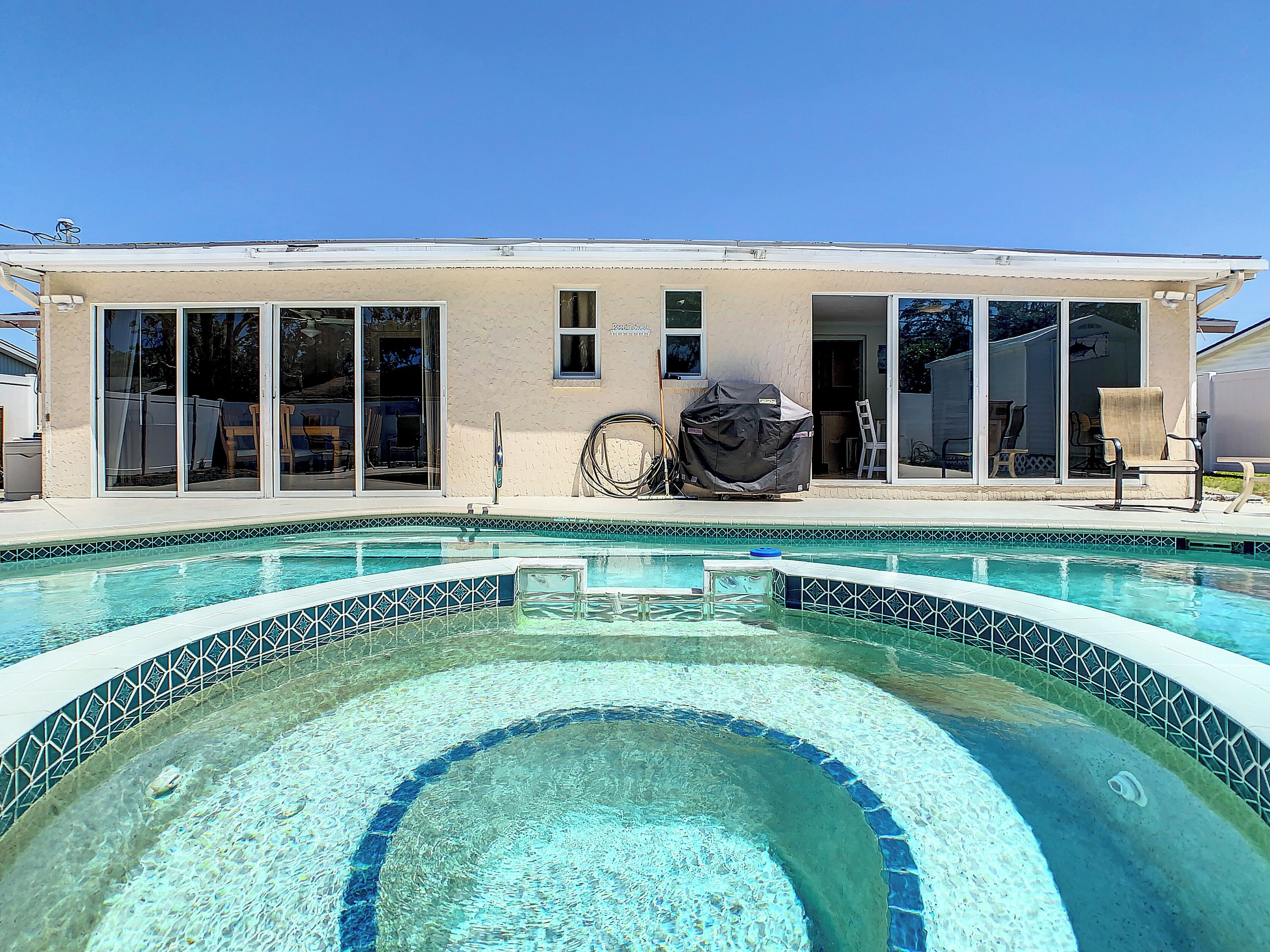 Une piscine résidentielle avec un bord carrelé, un barbecue couvert et une maison dotée de grandes portes-fenêtres coulissantes.