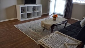 A living room with a dark sofa, a coffee table, a TV on a white stand, and a patterned rug.