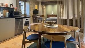 Een keuken met een wasmachine, een eettafel met stoelen, een televisie en een bed op de achtergrond.