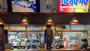A bar area with stools, a television displaying a parking lot, and a neon sign advertising '70s-80s music. - Peaky Blinder themed hotel rooms/studio apartments (Dudley)