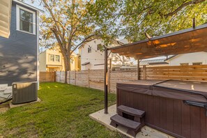 Relax in the backyard patio area with a hot tub, shaded by a pergola and surrounded by a lawn and a tree.