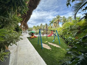 A tropical garden with a swing set, a treehouse, and a thatched-roof structure.