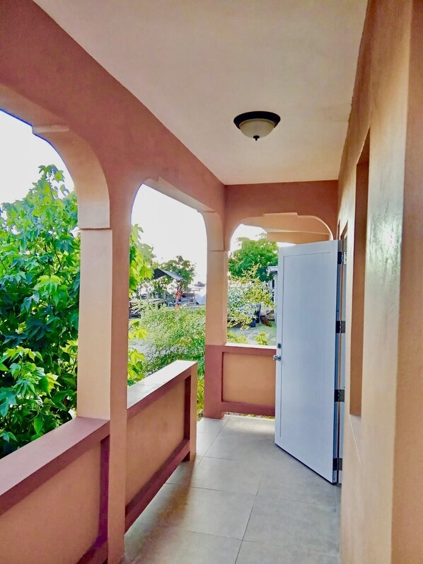A corridor with a white door, beige walls, and a view of greenery outside.