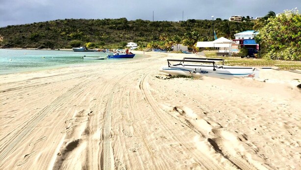 Une plage de sable avec des empreintes, un petit bateau et quelques bâtiments en arrière-plan.