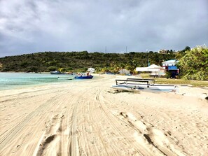 A sandy beach with footprints, a small boat, and a few buildings in the background.
