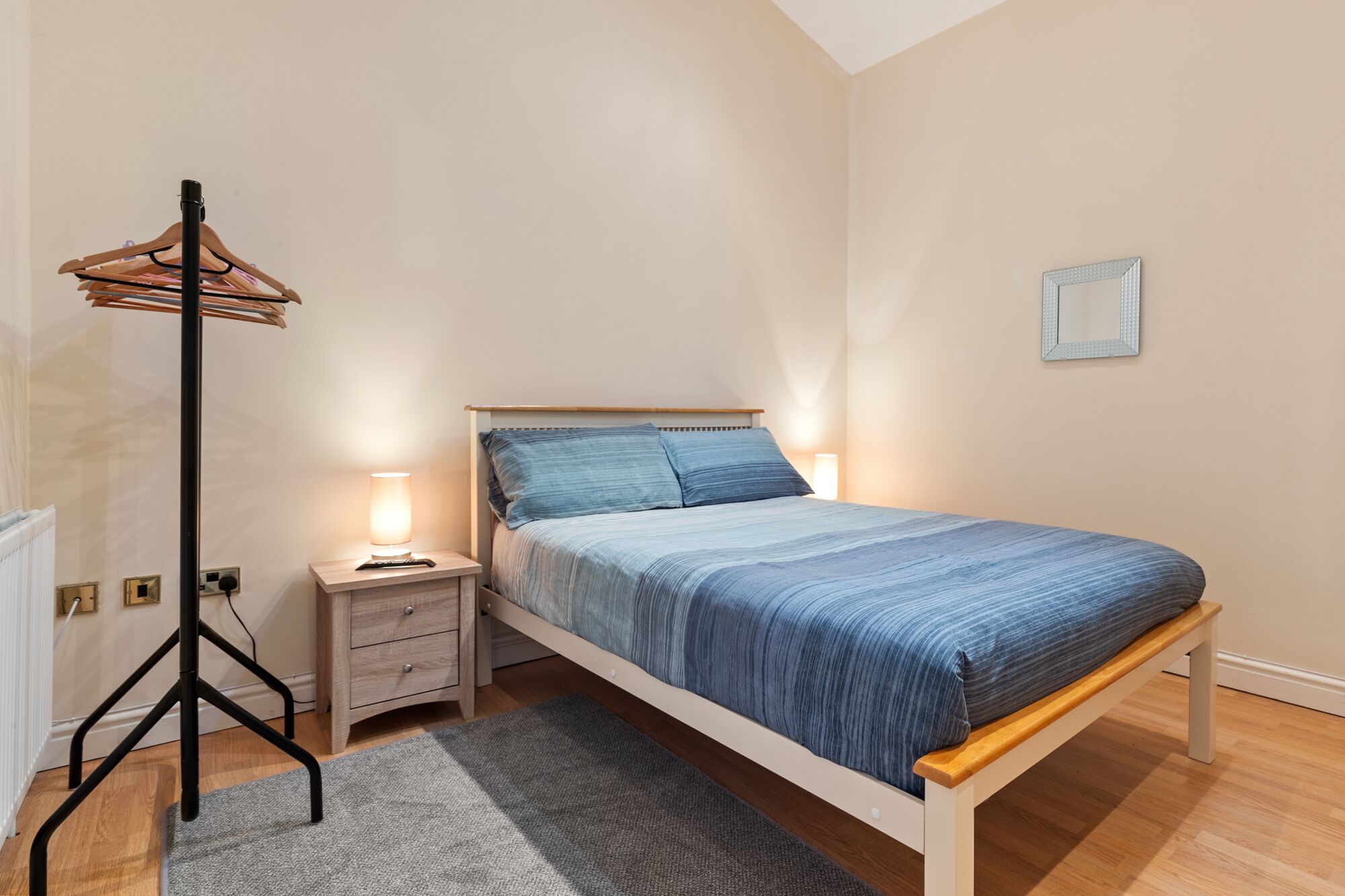 A bedroom with a bed, bedside tables, a floor lamp, a radiator, and a small wall-mounted picture.