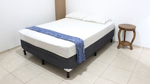 Una cama con una manta azul y almohadas blancas, un taburete de madera y una pared blanca lisa.