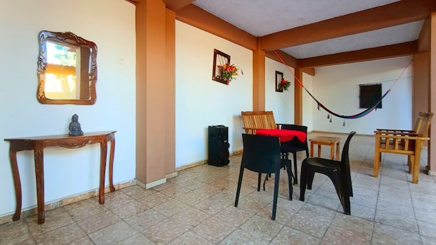 Una habitación con suelo de baldosas, una mesa de madera, dos sillas, una mesita con una estatua, una hamaca y un espejo adosado a la pared.