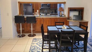 A compact kitchen with wooden cabinets, a microwave, and a coffee maker. There is a dining table with chairs and a laptop on a small desk.