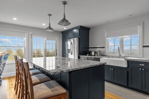 Une cuisine moderne avec des meubles de couleur sombre, un réfrigérateur en acier inoxydable, un grand îlot central doté de sièges et une vue sur l’extérieur grâce à de grandes fenêtres.