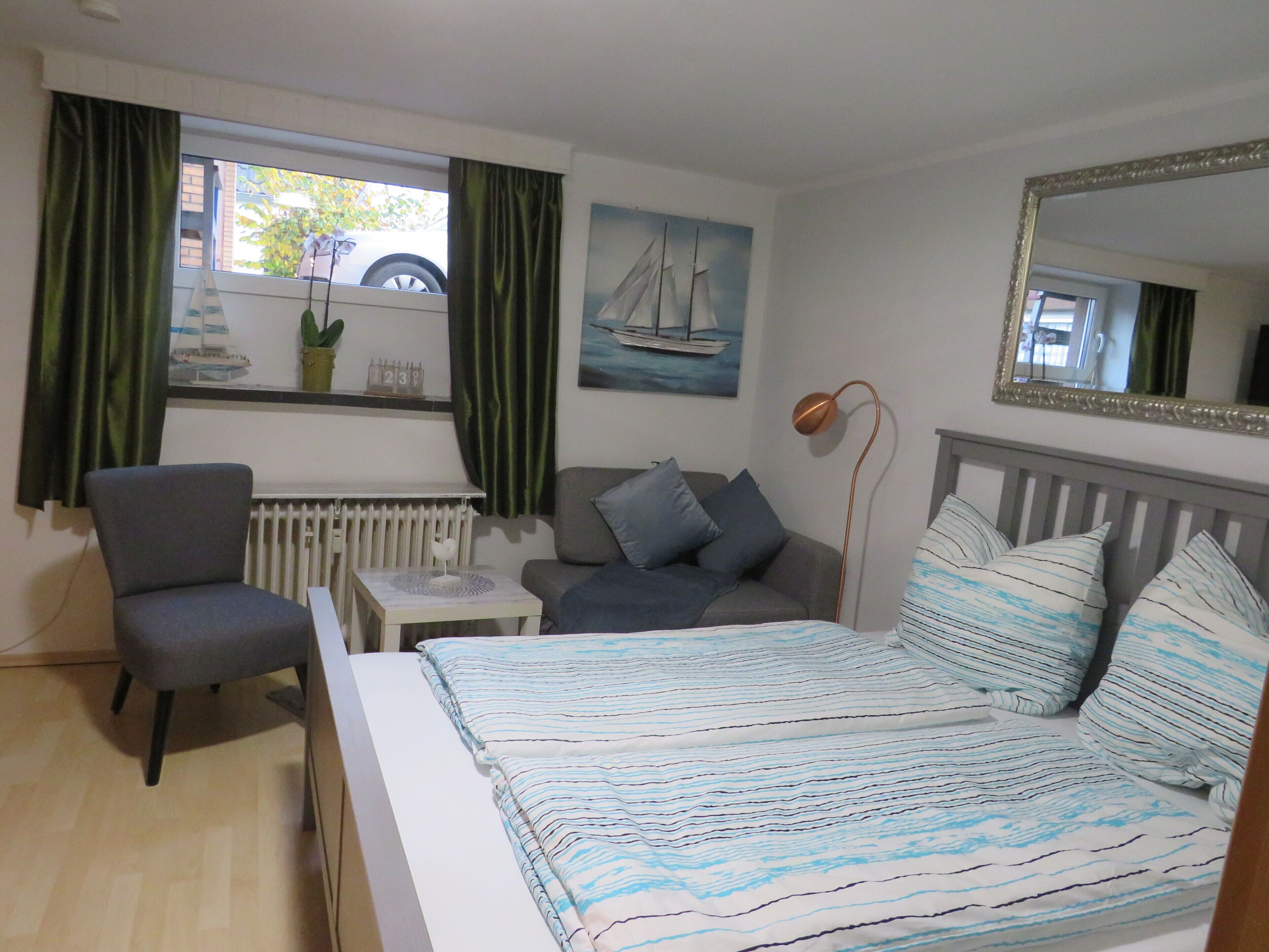 Ein Schlafzimmer mit Bett, Sessel, Sofa, einem kleinen Tisch, einer Heizung, einem Fenster mit Vorhängen, einem Gemälde und einem Spiegel.