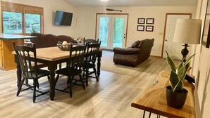 A dining area with a wooden table and chairs, a sofa, a TV mounted on the wall, and a ceiling fan. - Riverfront House near Cow Shoals - King bed & WiFi (Heber Springs)