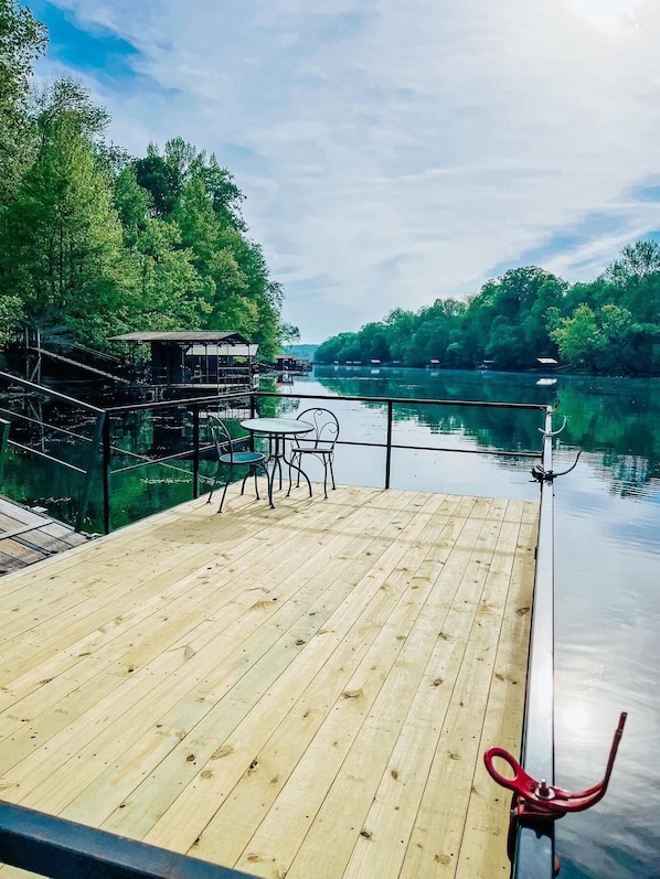 Eine Holzterrasse mit Tischen und Stühlen bietet Blick auf einen ruhigen Fluss, umgeben von üppigem Grün.