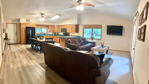 A spacious living area with a dark brown sofa, a dining table with chairs, a kitchen area with wooden cabinets, and a ceiling fan. - Riverfront House near Cow Shoals - King bed & WiFi (Heber Springs)