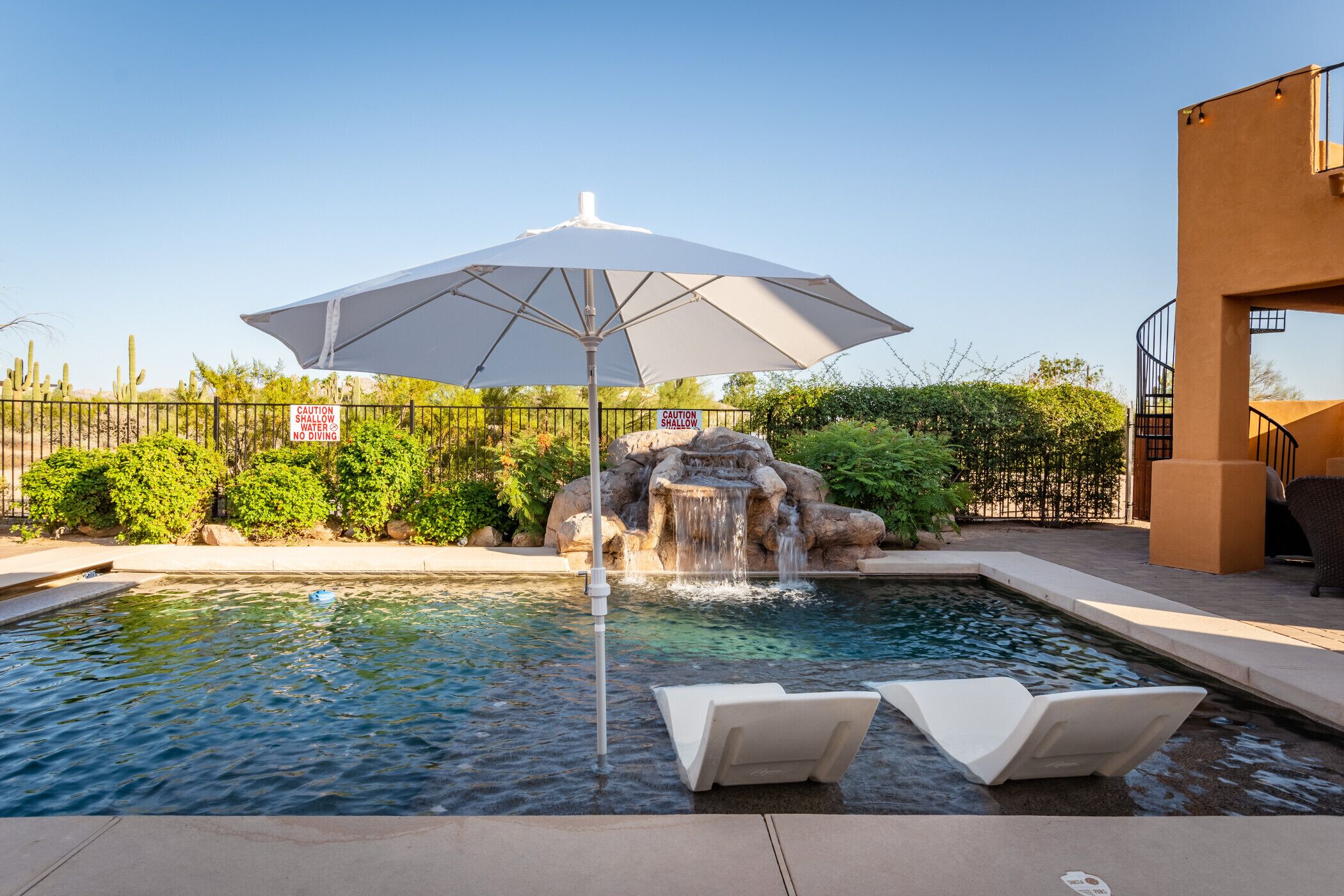 A swimming pool with a waterfall feature, surrounded by a fence, and an umbrella providing shade.