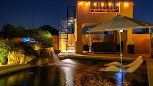 A nighttime view of a pool area with a waterfall, a covered seating area, and a building with outdoor lighting. - Private Desert Oasis with Pool & Hot Tub, Game Room | Family & Pet Friendly (Scottsdale)