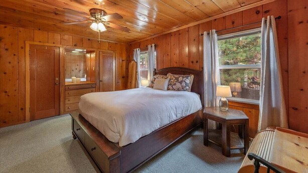 A wooden cabin bedroom with a bed, a nightstand, a window with curtains, and a ceiling fan.