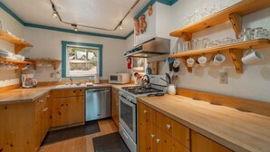 A compact kitchen with wooden cabinets, a stove, a microwave, and a sink. There are shelves with glassware and a window with a view.