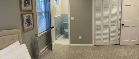 A bedroom with a bed, a window with blinds, a door leading to a bathroom, and a closet with louvered doors.