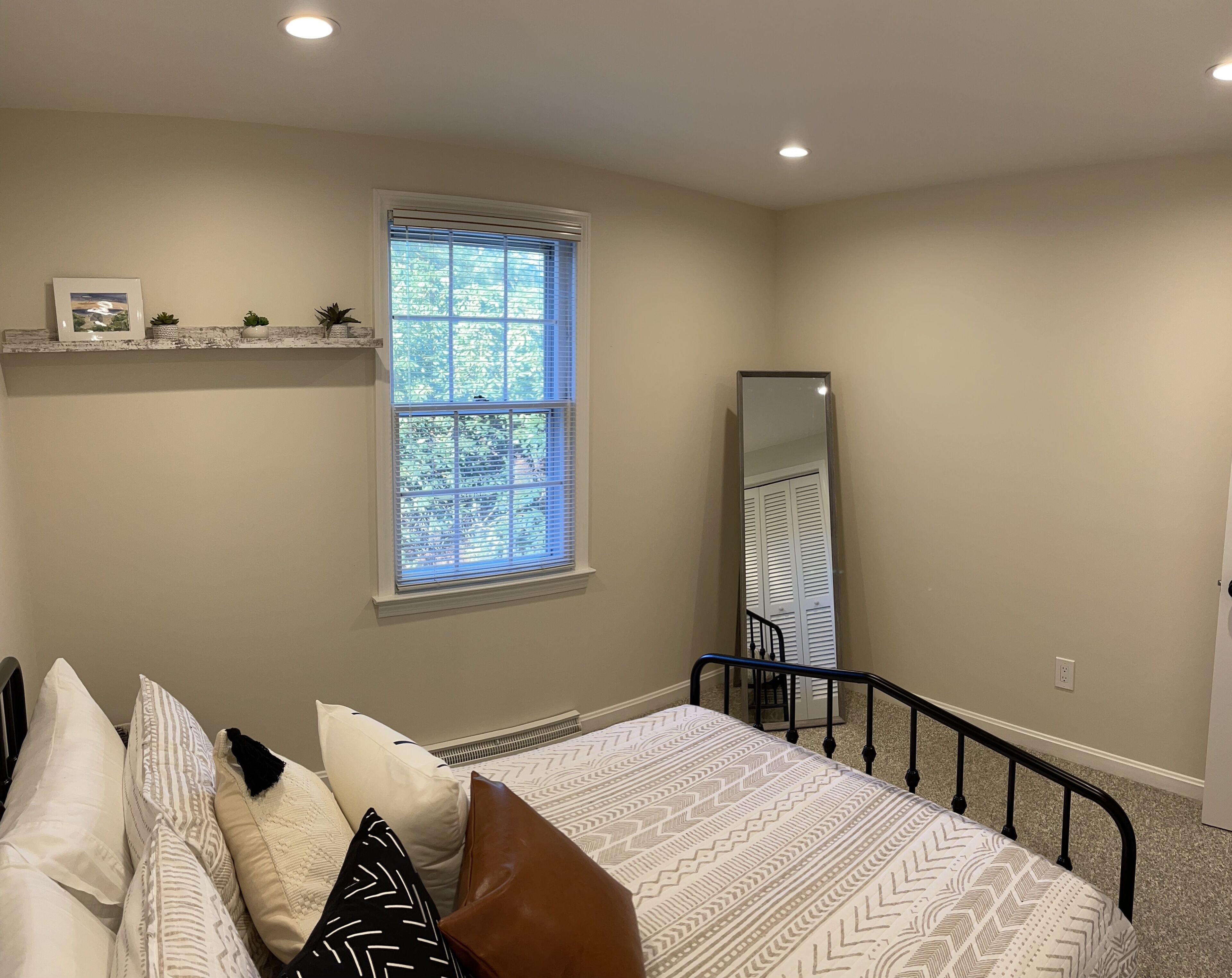 A bedroom with a bed, pillows, a window, a mirror, and a shelf with decorative items.