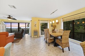 Un coin repas avec une table en bois et des chaises en osier, un ventilateur de plafond et une vue sur le coucher de soleil à travers de grandes fenêtres.