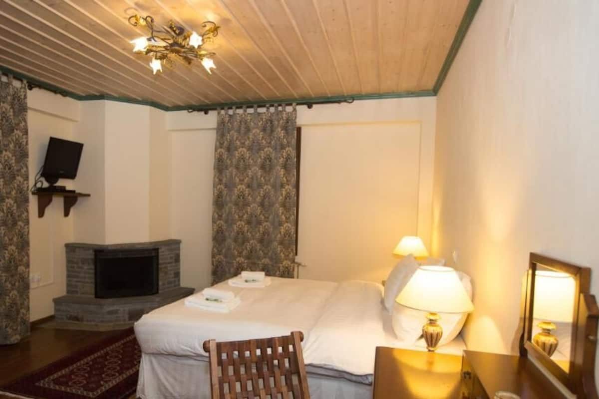A hotel room with a fireplace, a bed with two pillows, a nightstand with a lamp, and a wooden chair.