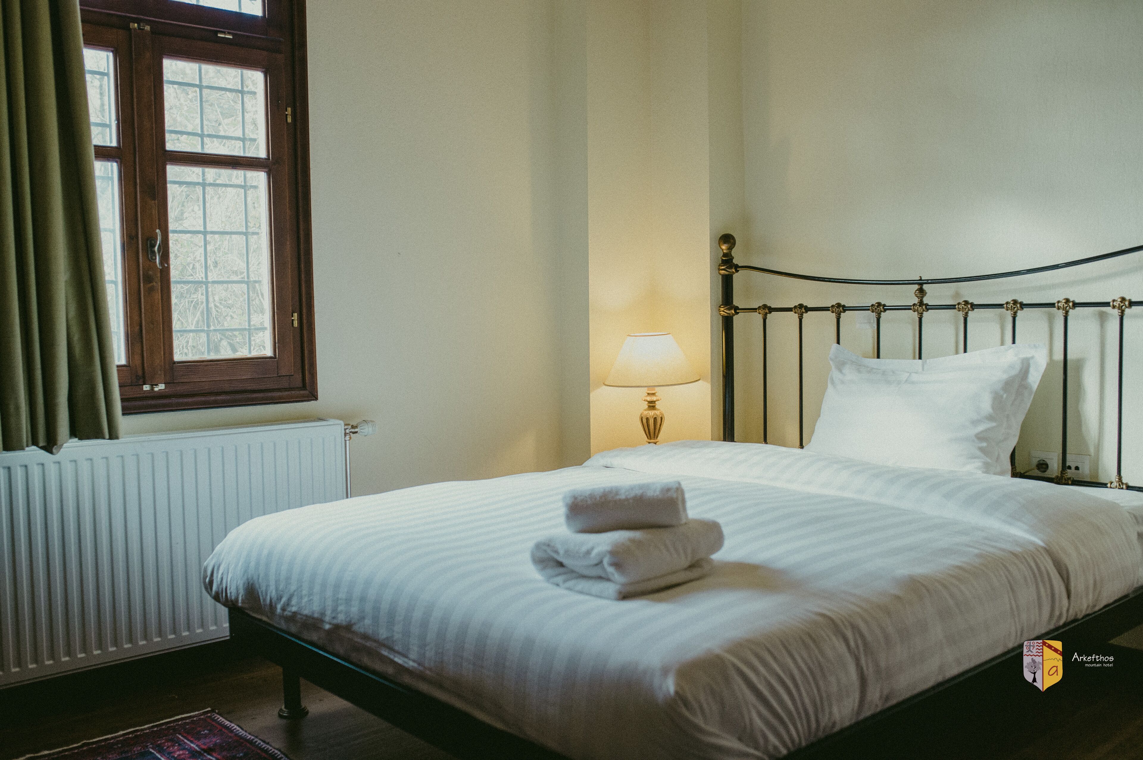 Una cama bien hecha con sábanas blancas y toallas dobladas, una lámpara de noche y una ventana con cortinas.