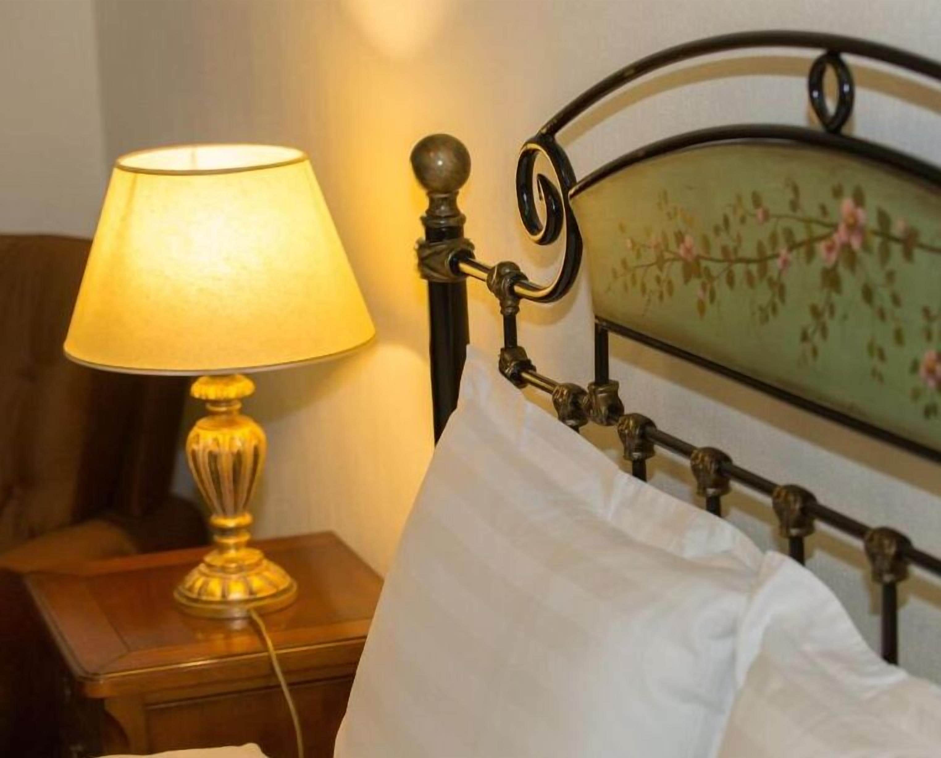 Un lit avec un oreiller blanc, une tête de lit en laiton et une lampe de table avec un abat-jour jaune.