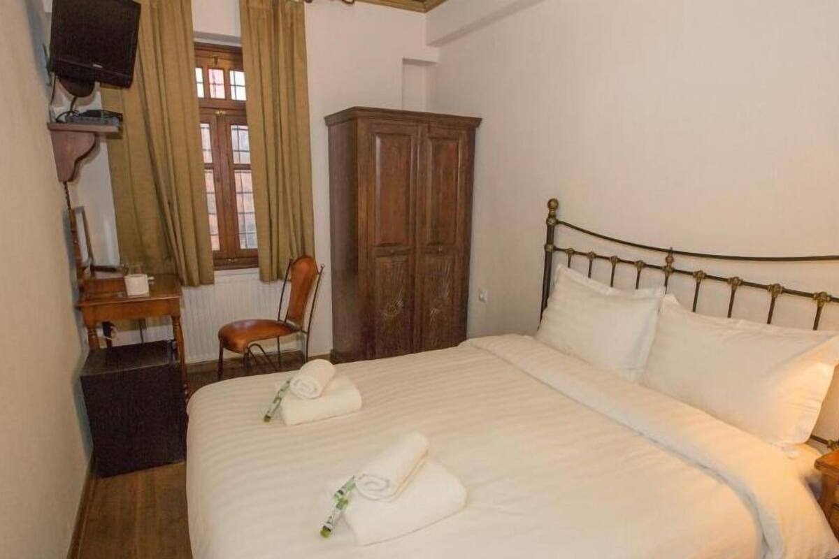 A bedroom with a bed, a wooden wardrobe, a chair, a desk, and a television.