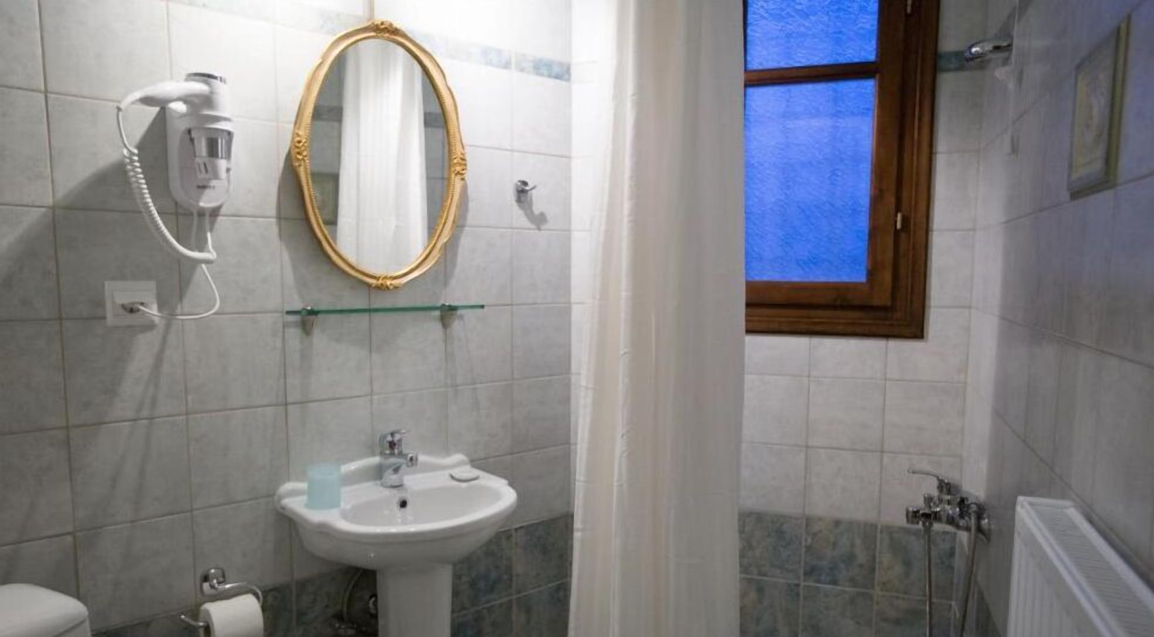 Une salle de bain avec un lavabo blanc, un miroir à cadre doré, un sèche-cheveux et une fenêtre avec du verre teinté en bleu.