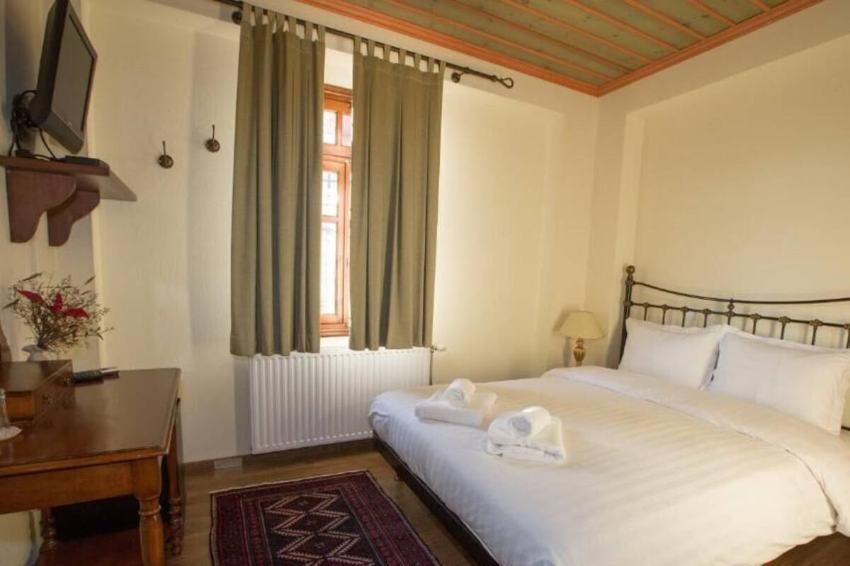 A bedroom with a bed, a bedside table, a lamp, a window with curtains, a radiator, and a television on a stand.