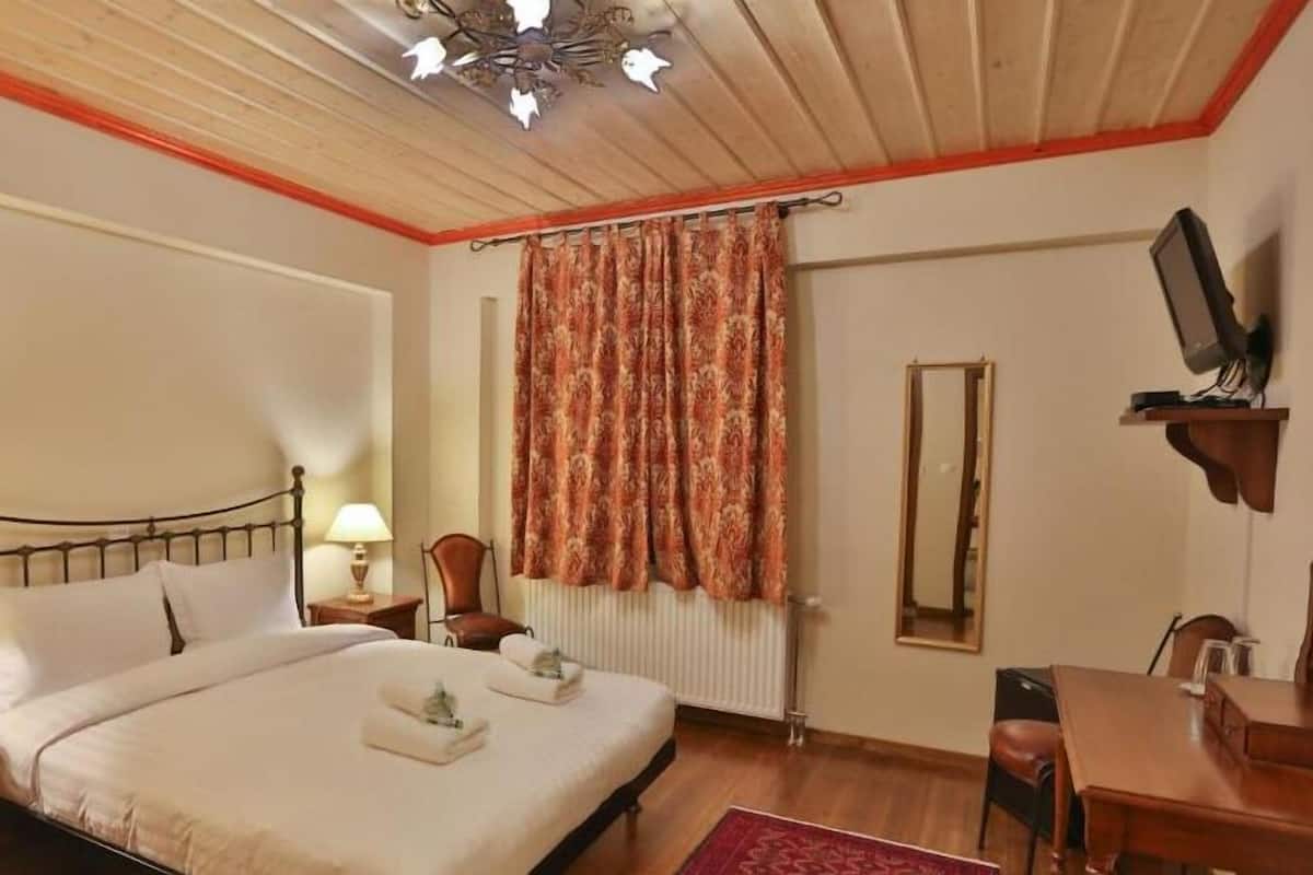 A bedroom with a bed, two bedside tables with lamps, a chair, a desk with a computer, a mirror, and a window with curtains.