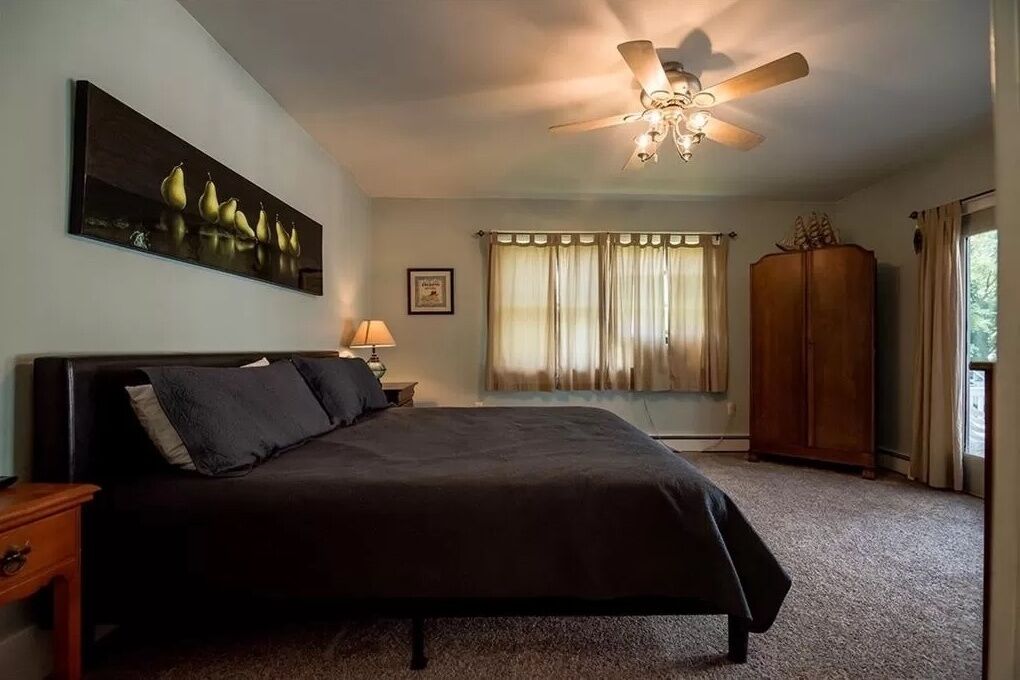 Un dormitorio con cama, ventilador de techo, ventana con cortinas, mesita de noche y armario.