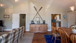 A cozy living room with a couch, a dining table with chairs, a kitchen area with a wooden cabinet, and a hallway with a door. - Keuka East: Spacious & Comfy (Penn Yan)