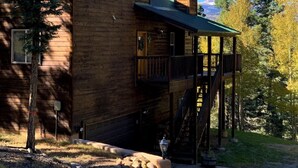 Una cabaña de madera con techo verde, una chimenea y una terraza con escaleras que llevan a un segundo nivel.