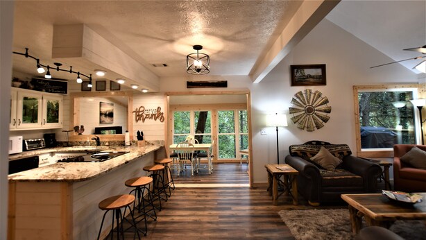 Un'ampia cucina con angolo bar, sgabelli da bar in legno e un tavolo da pranzo con sedie. È presente un'ampia finestra con vista sugli alberi, una ventola a soffitto e un elemento decorativo a parete.