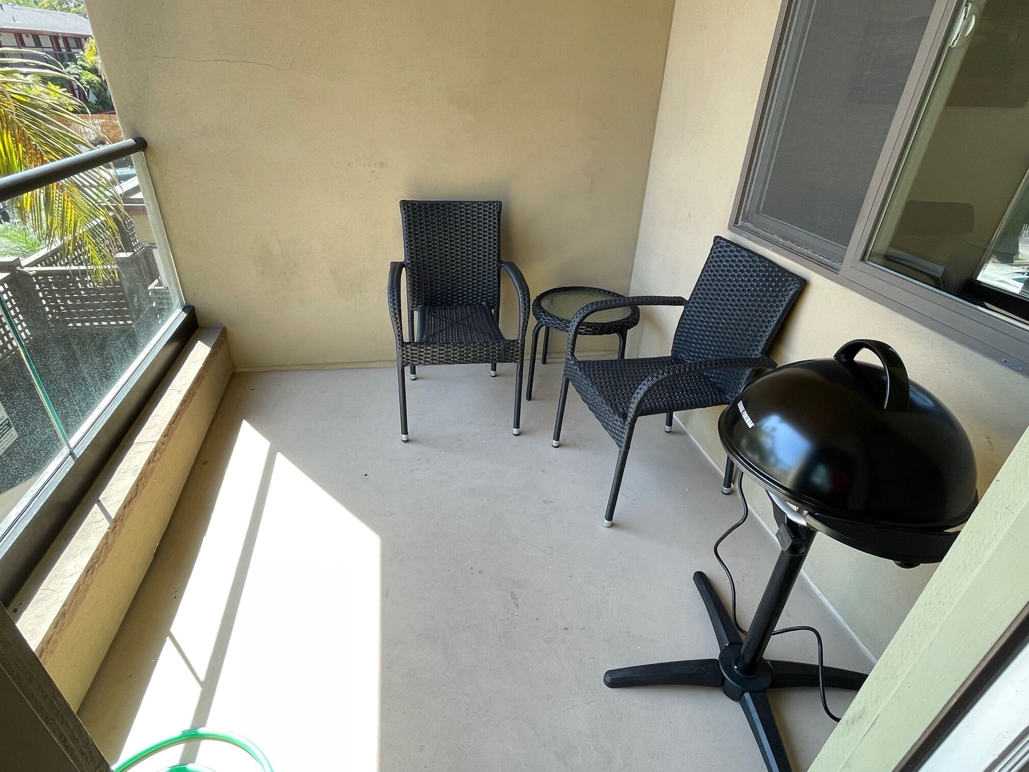 A balcony with two wicker chairs and a small table, a grill, and a window overlooking a garden.