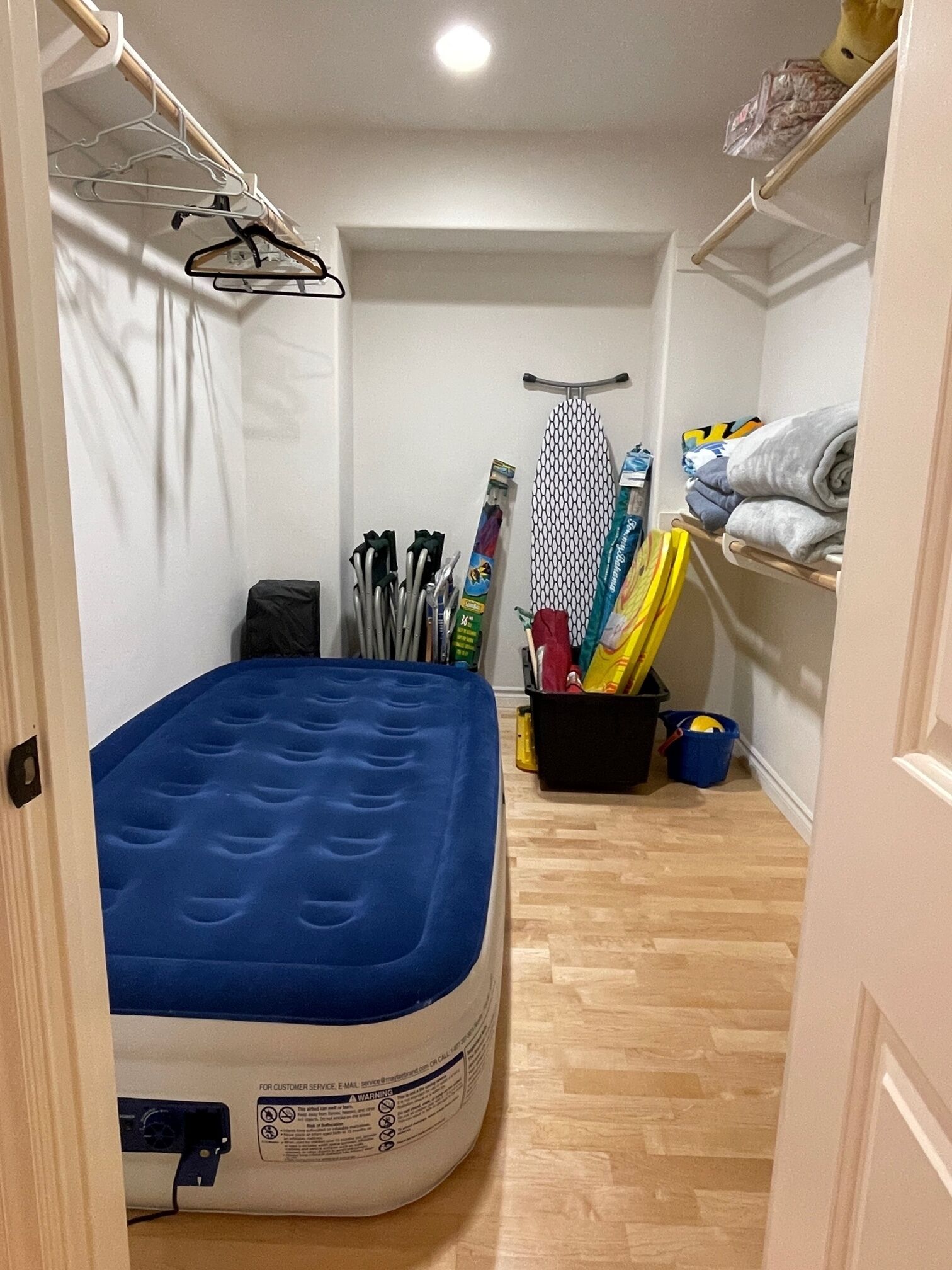 Ein kleiner, schmaler Raum mit einer blauen Matratze, einer weißen Wand, einem Holzboden, einem Kleiderständer, einem Regal mit gefalteter Kleidung und einem Regal mit Reinigungsmitteln.