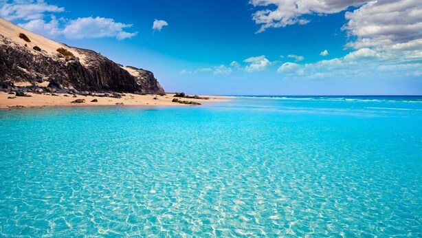 Un mar turquesa transparente con una playa arenosa y acantilados rocosos bajo un cielo azul.