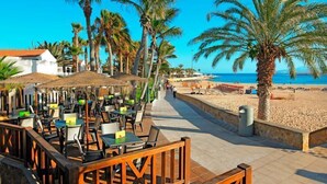 Un área de comedor al aire libre con mesas y sillas, palmeras y una playa al fondo.