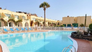 Una piscina con agua azul, rodeada por un deck blanco. Hay sillas reclinables azules y una palmera.