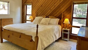 Um quarto com teto inclinado de madeira, estrutura de cama de madeira, duas mesas de cabeceira com abajures e vista para árvores pelas janelas.