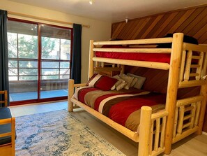 Un lit superposé avec une couvre-lit rouge et beige, une tête de lit en bois et une vue sur un balcon doté d’une balustrade en bois.