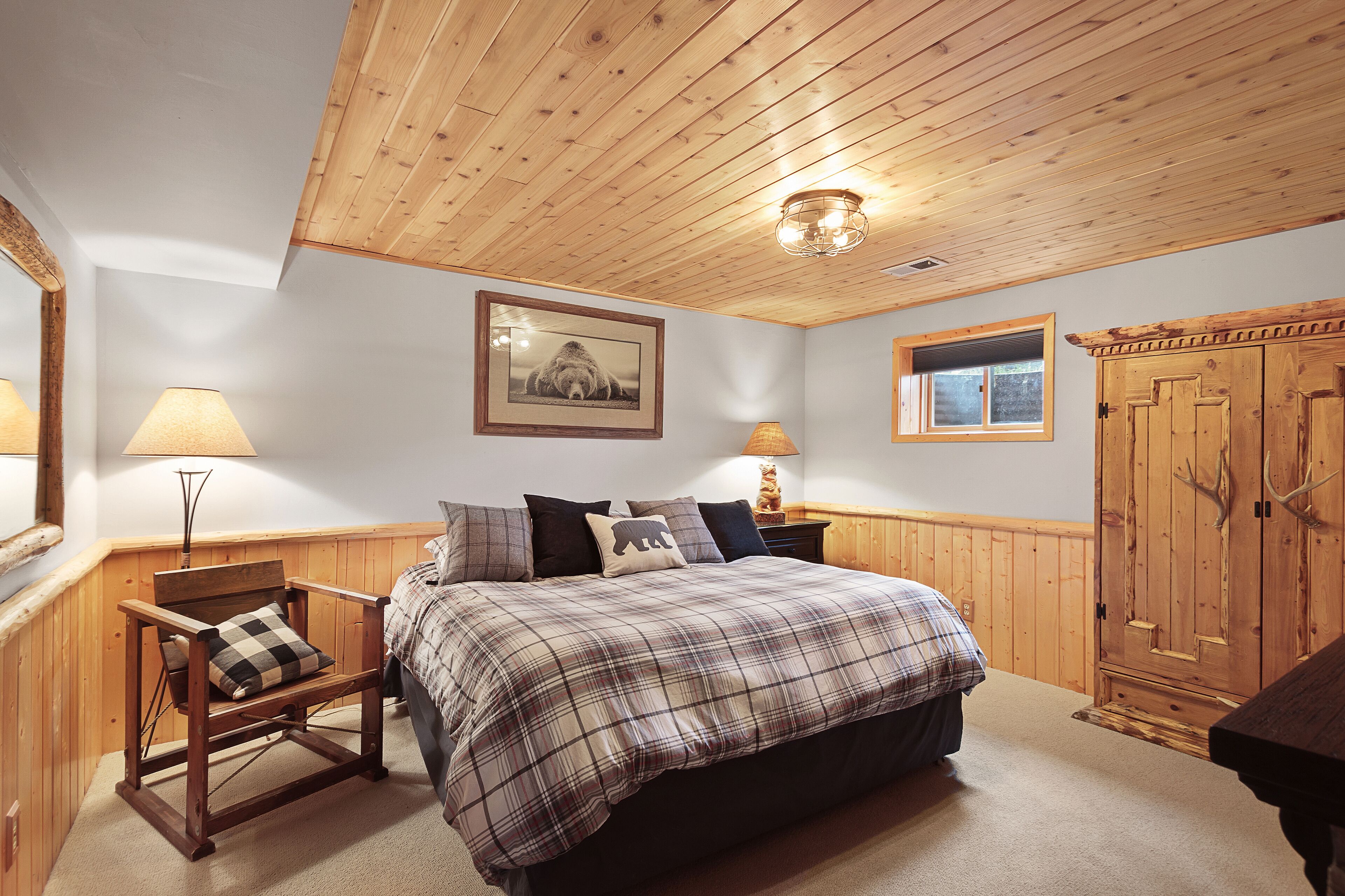 A bedroom with a wooden ceiling, a bed with a plaid bedspread, two bedside lamps, a wooden wardrobe, and a framed picture on the wall.