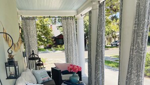 Un área de estar al aire libre techada con mobiliario de mimbre, un jarrón decorativo con flores y cortinas estampadas.