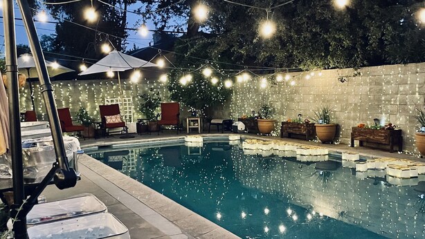 Una piscina con pared de piedra, luces colgantes y plantas en macetas.