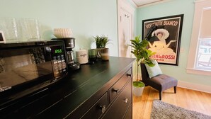 Una cocina con microondas, cafetera y una taza de café sobre el mostrador. Hay una planta en el suelo y un cartel en la pared.