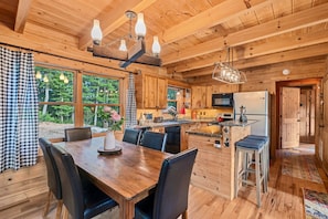 Interior de una cabaña de madera con comedor, cocina y grandes ventanales.