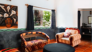 Una habitación con un cuadro grande, dos sillones de mimbre, una mesa redonda y una ventana con cortinas.