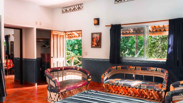 Una habitación con una cama, dos sillones de mimbre, un sofá, una ventana con cortinas y vistas al exterior.